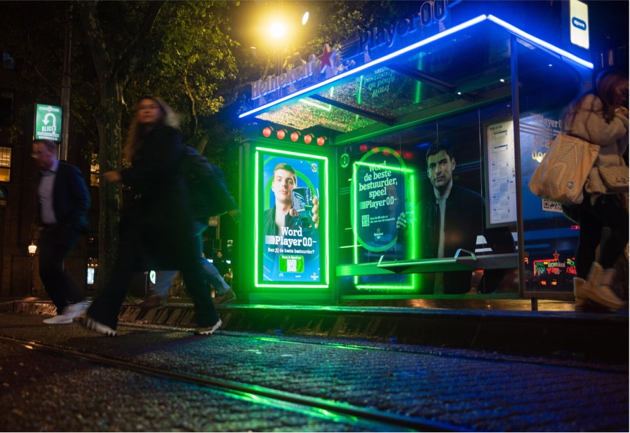 A Player 0.0 branded city tram shelter at night, its screen inviting passers-by to play
