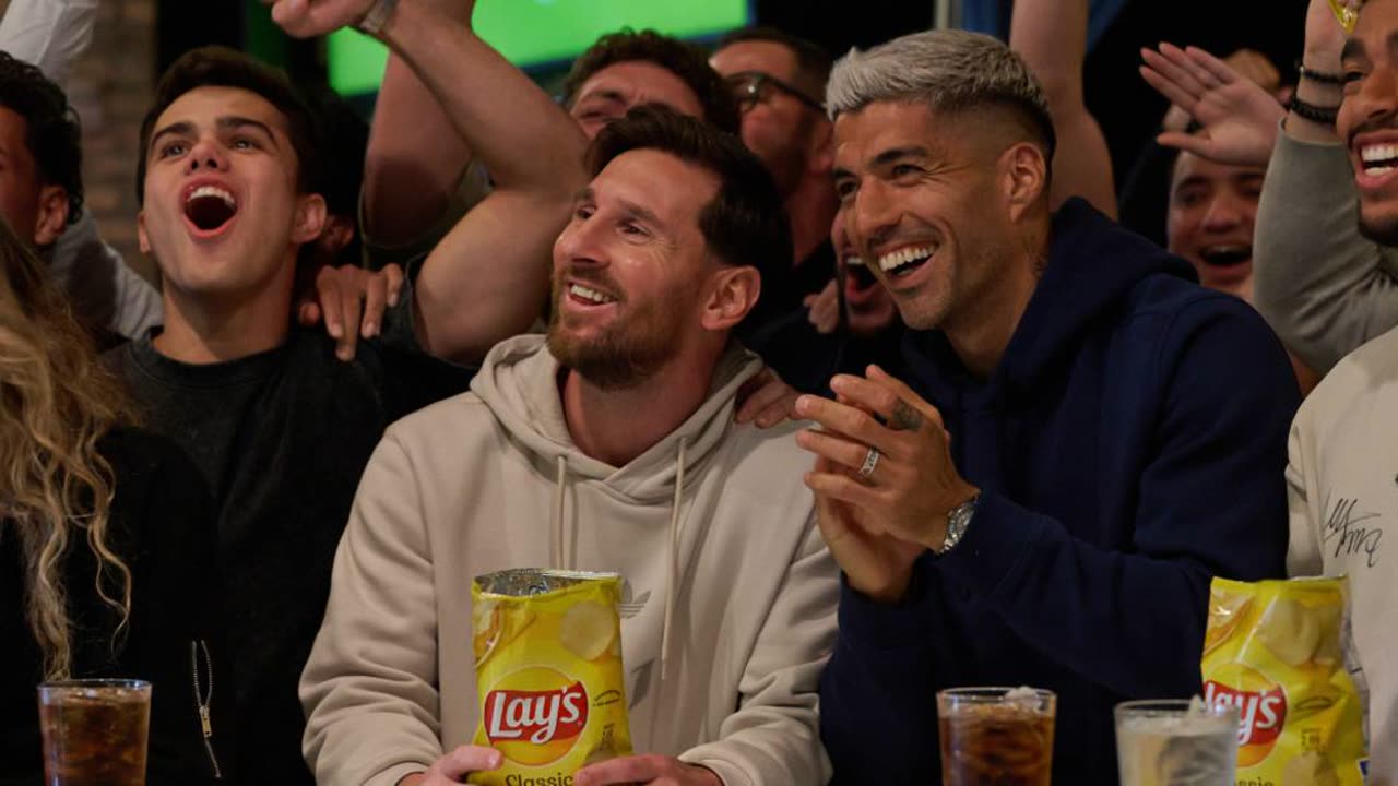 Lionel Messi and Luis Suárez among cheering fans, each holding a bag of Lay’s while watching a match